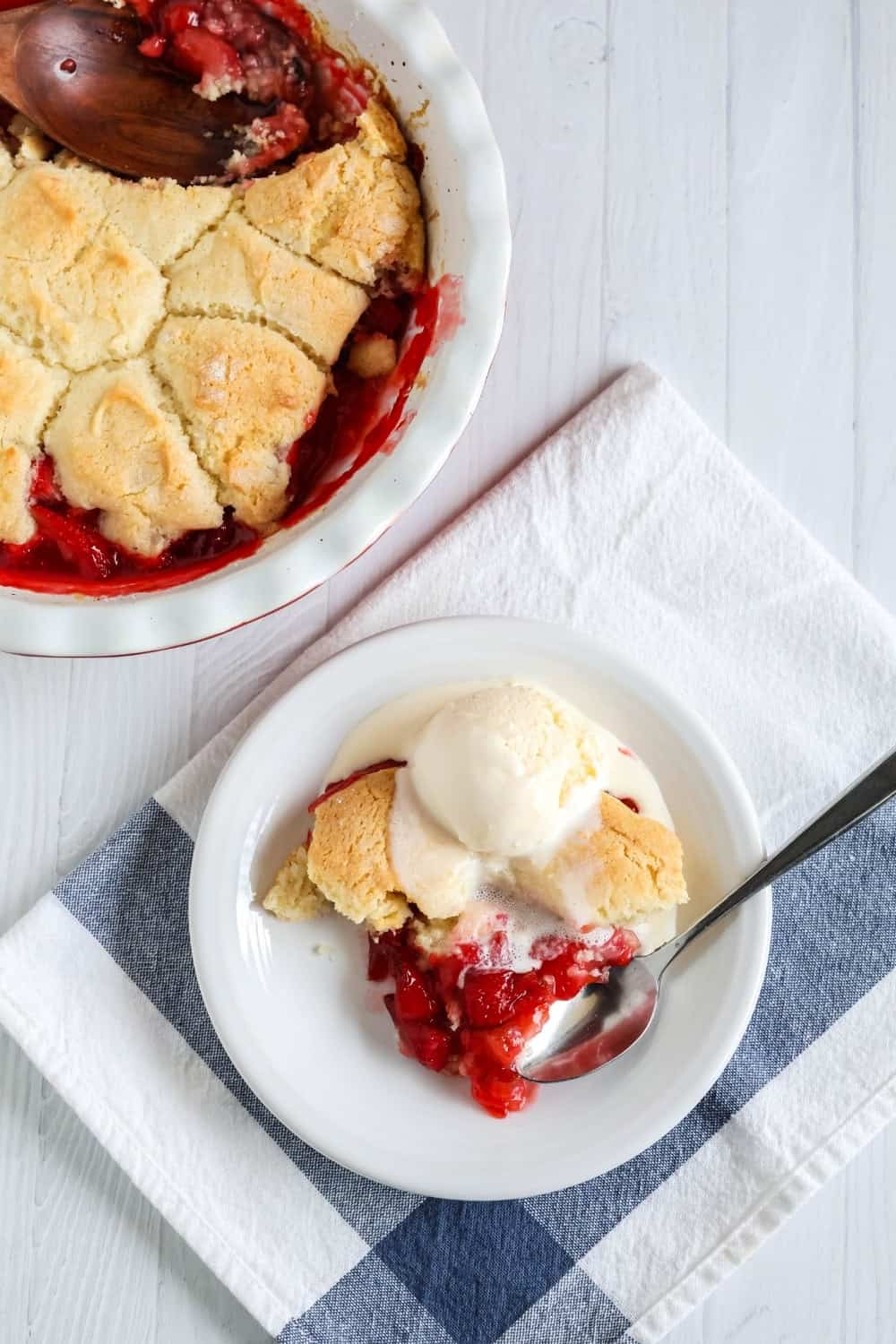 overhead view of a white plate with a serving of strawberry cobbler made with Bisquick, topped with ice cream. Next to the plate is a deep dish pie pan of the remaining cobbler.