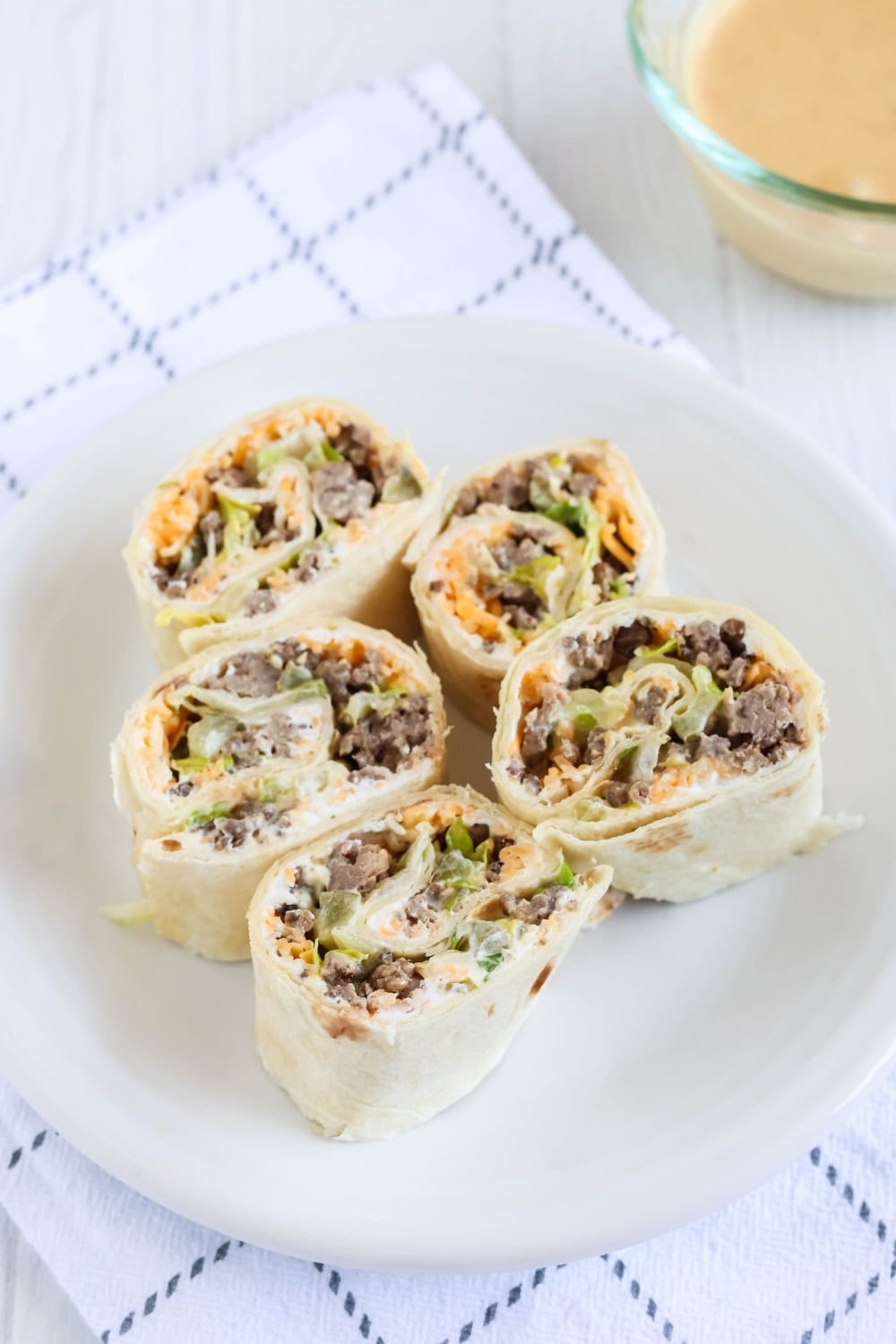 a white plate with five cheeseburger pinwheels on it, along with dipping sauce in the background.