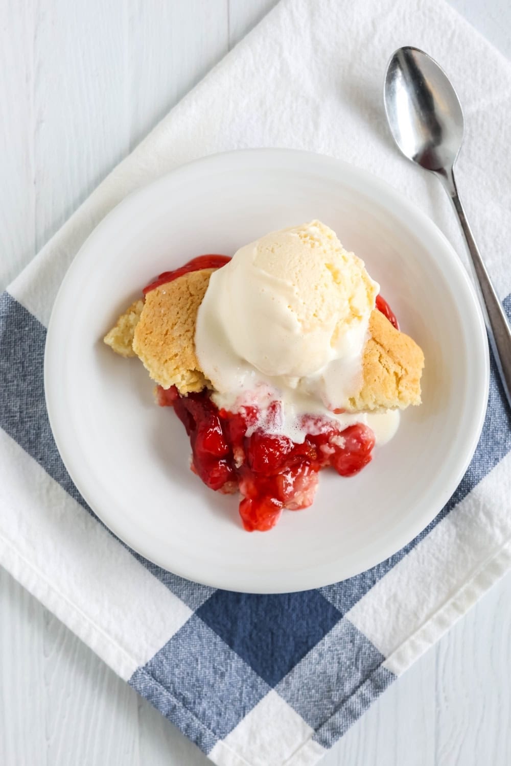 overhead view of Bisquick cobbler made with strawberries, served on a white plate, topped with vanilla ice cream. The plate is on top of a white and blue kitchen towel, with a spoon next to it.