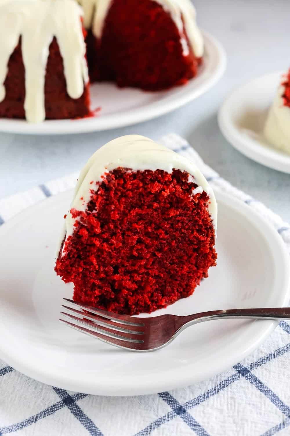 upright slice of red velvet bundt cake, topped with cream cheese frosting, served on a white plate, with the remaining cake in the background.
