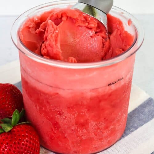 Ninja Creami strawberry sorbet in a pint container, with an ice cream scoop in it. Fresh strawberries are next to the container.