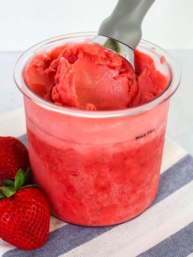 Ninja Creami strawberry sorbet in a pint container, with an ice cream scoop in it. Fresh strawberries are next to the container.