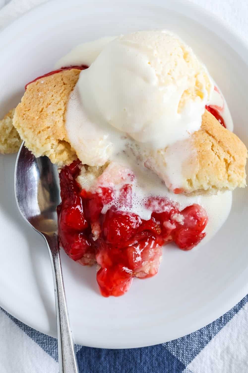 warm fresh strawberry Bisquick cobbler, topped with a scoop of melting vanilla ice cream, served on a white plate with a spoon.