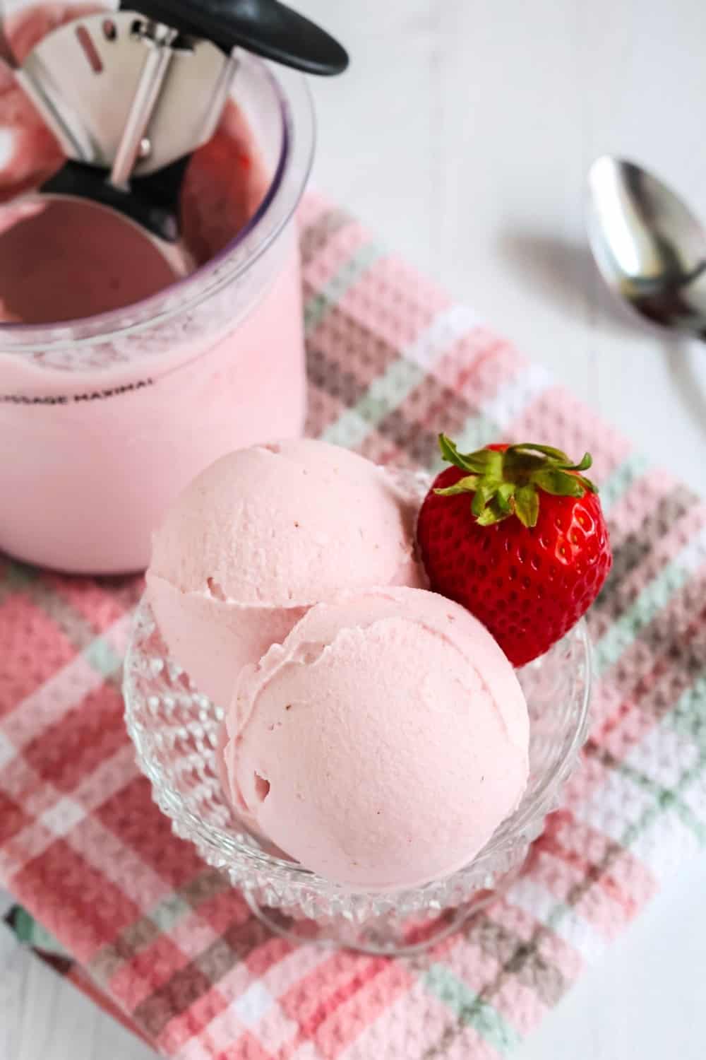 scoops of strawberry gelato in a glass dish with a fresh strawberry. The Ninja Creami pint container of remaining gelato is in the background, along with a spoon.