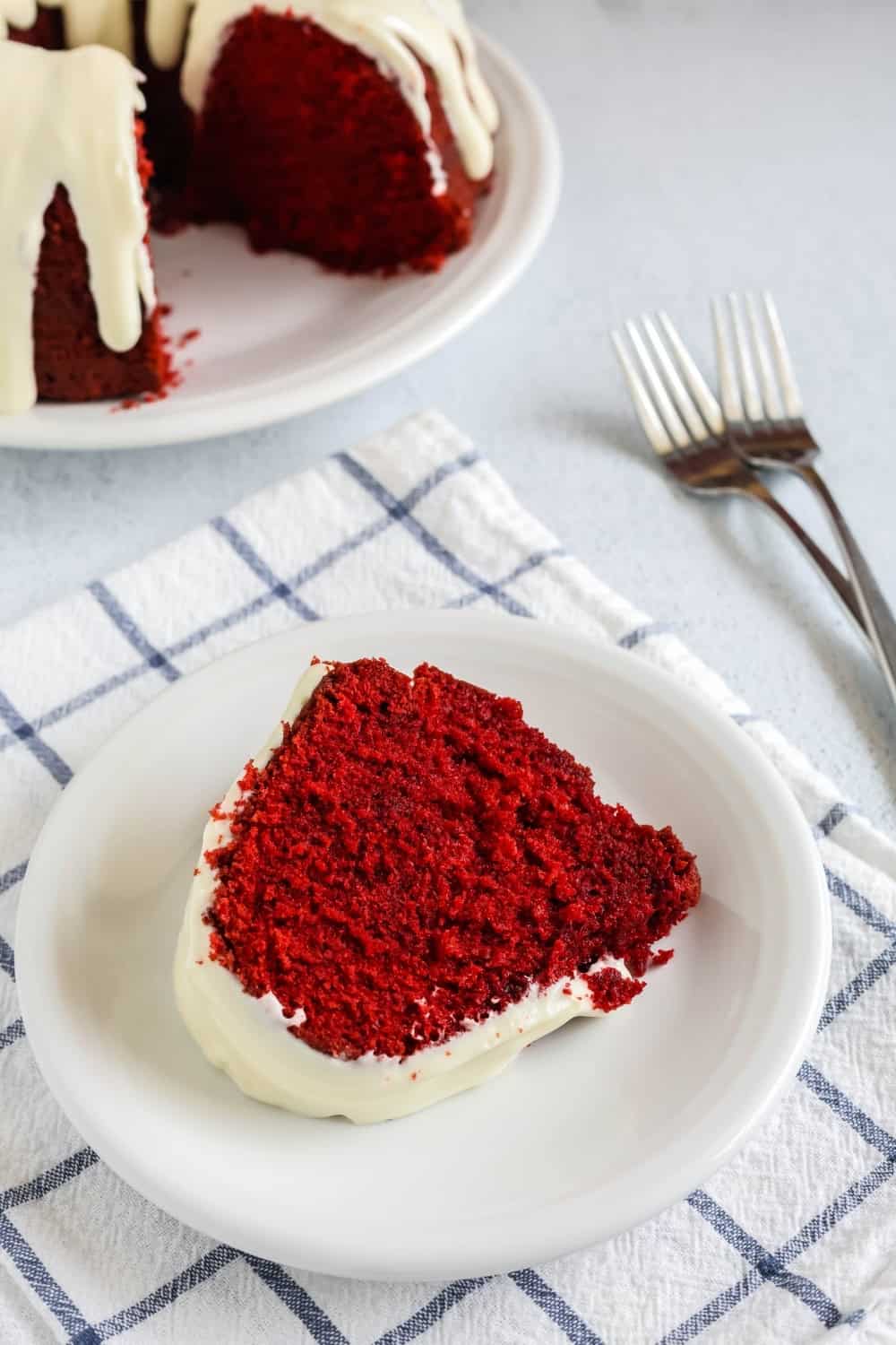 slice of red velvet cake, with cream cheese icing, lying on its side on a white plate. Two forks and the remaining bundt cake are in the background.