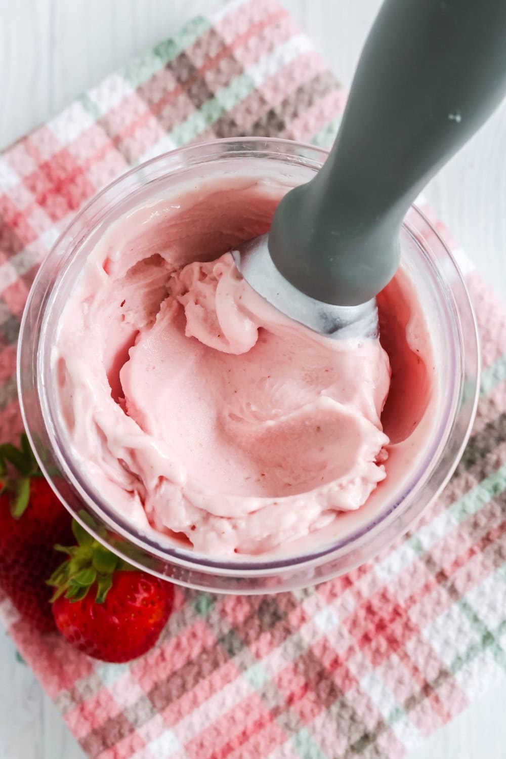 overhead view of a pint of rich and creamy strawberry gelato made in the Ninja Creami machine. An ice cream scoop is in the pint, and there are fresh strawberries near it.