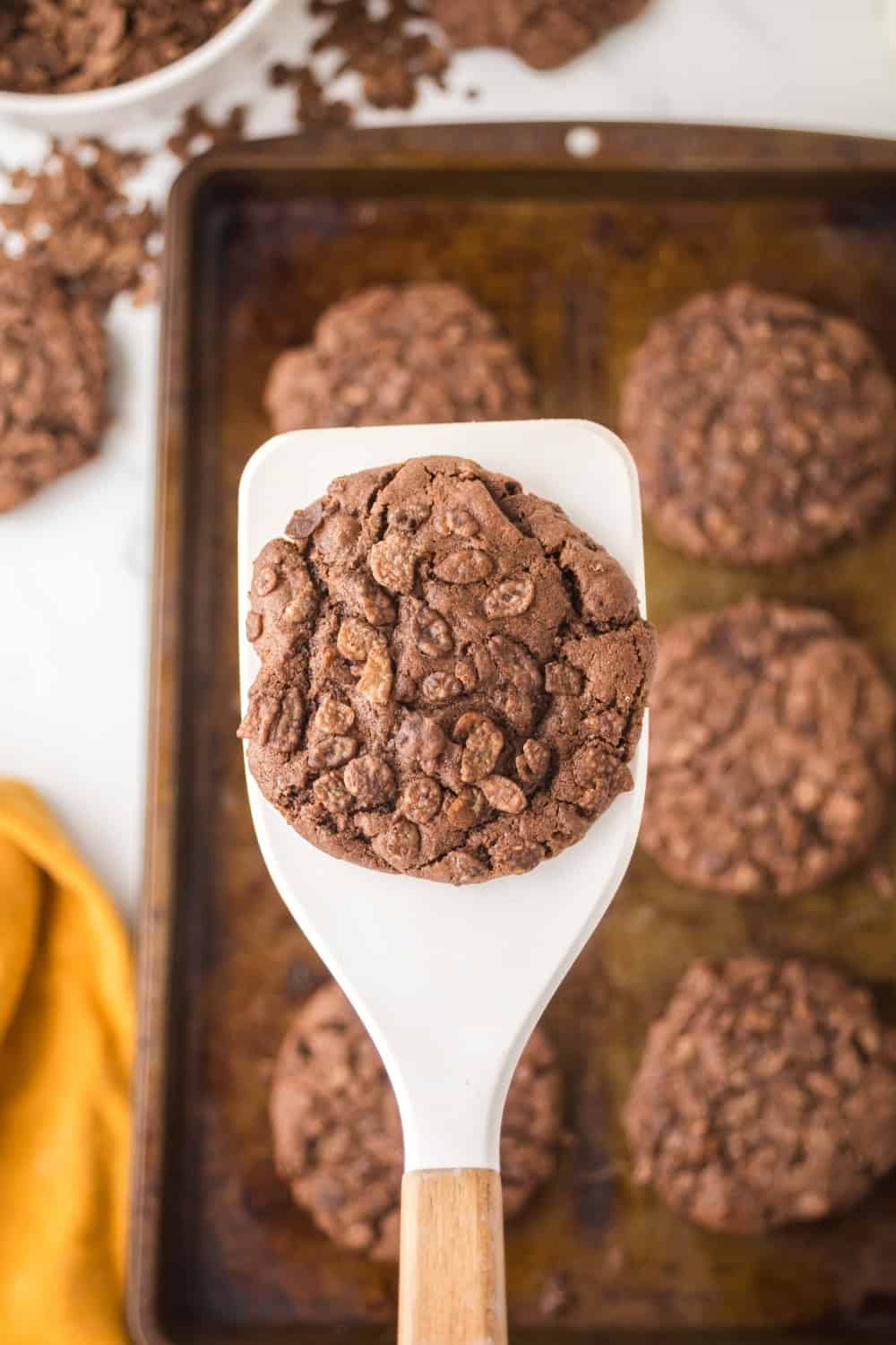 a white spatula lifts a cookie made with Cocoa Pebbles cereal off a baking sheet.
