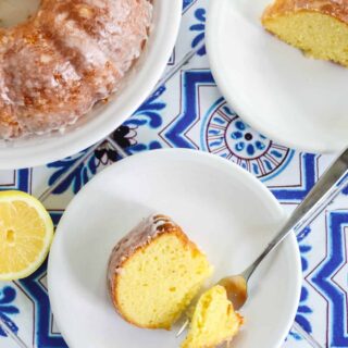 overhead view of a slice of lemon mayo bundt cake on a white plate, with a bite cut out by a fork. The bundt cake and another slice are in the background.
