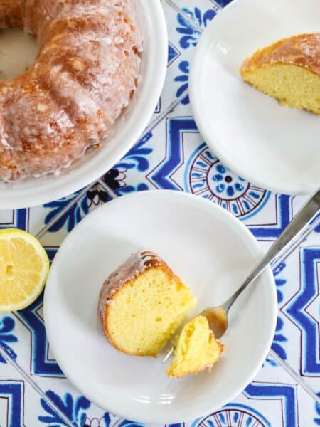 overhead view of a slice of lemon mayo bundt cake on a white plate, with a bite cut out by a fork. The bundt cake and another slice are in the background.