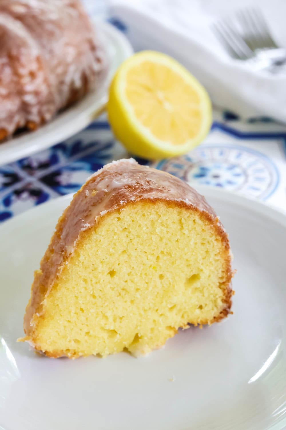 close-up view of a slice of Duke's lemon mayo cake on a white plate. A lemon half is in the background, along with forks and the bundt cake.