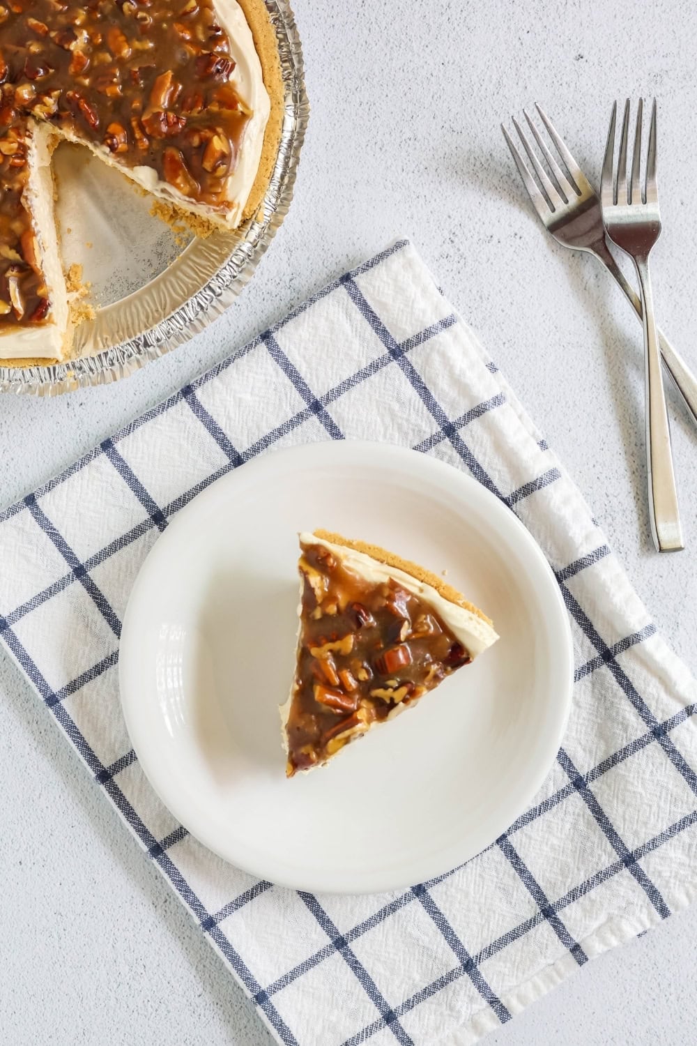 overhead view of a slice of pecan pie cheesecake on a white plate. The remainder of the cheesecake is in the background, along with two forks.