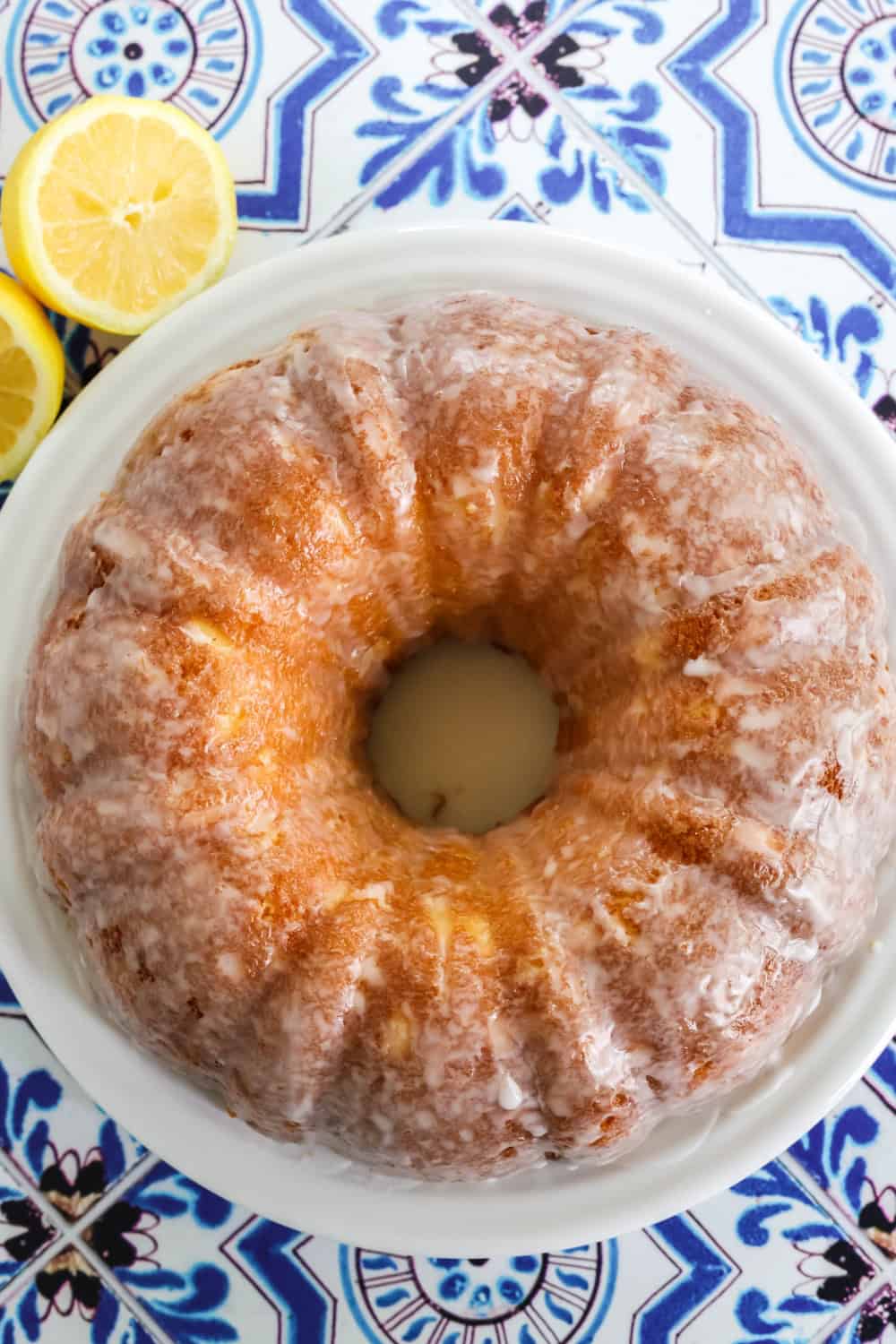 overhead view of a lemon mayonnaise bundt cake on a white platter, with two lemon halves next to the plate.