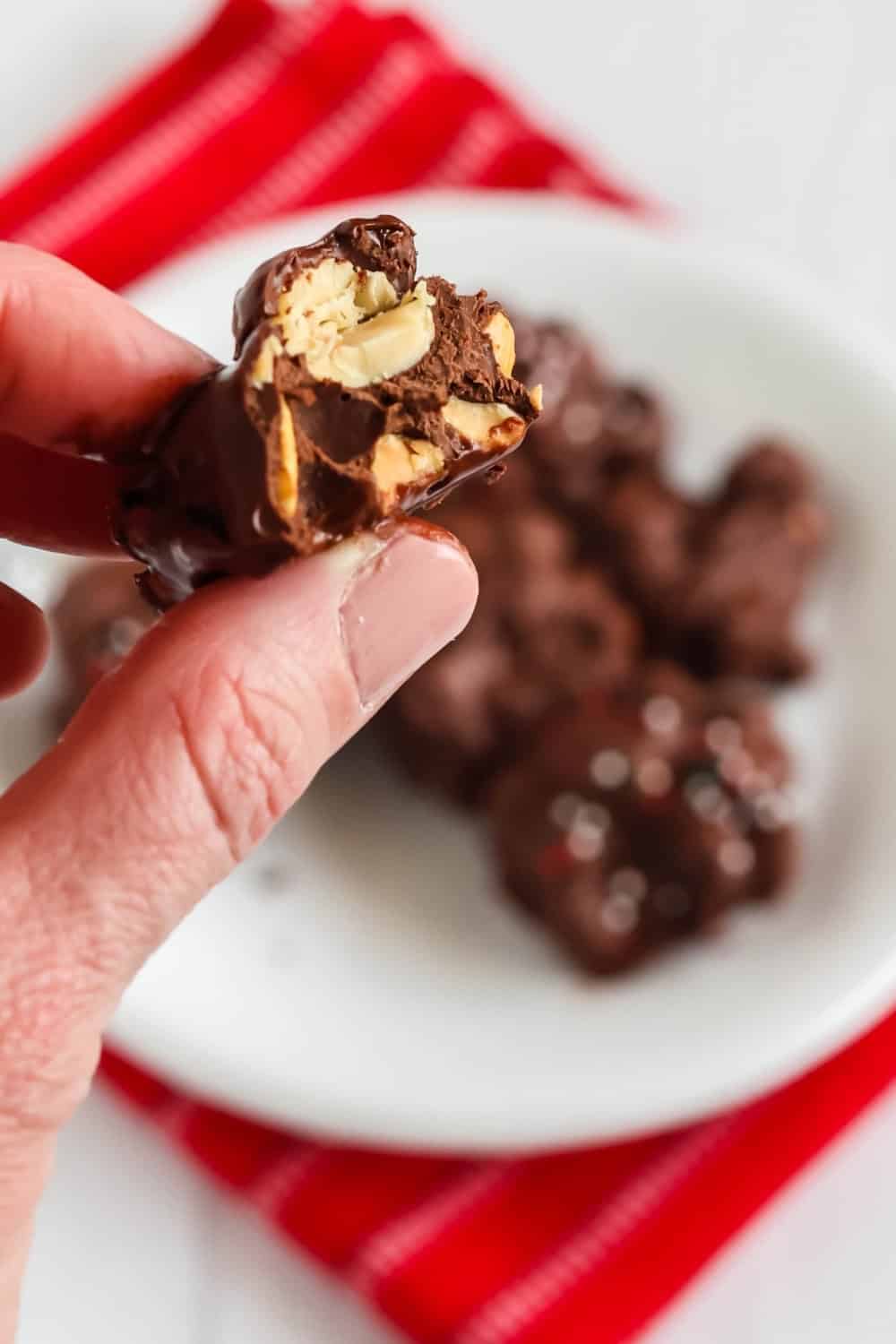 a hand holds a chocolate peanut cluster that has been bitten in half, showing the peanuts inside of the sugar-free chocolate.