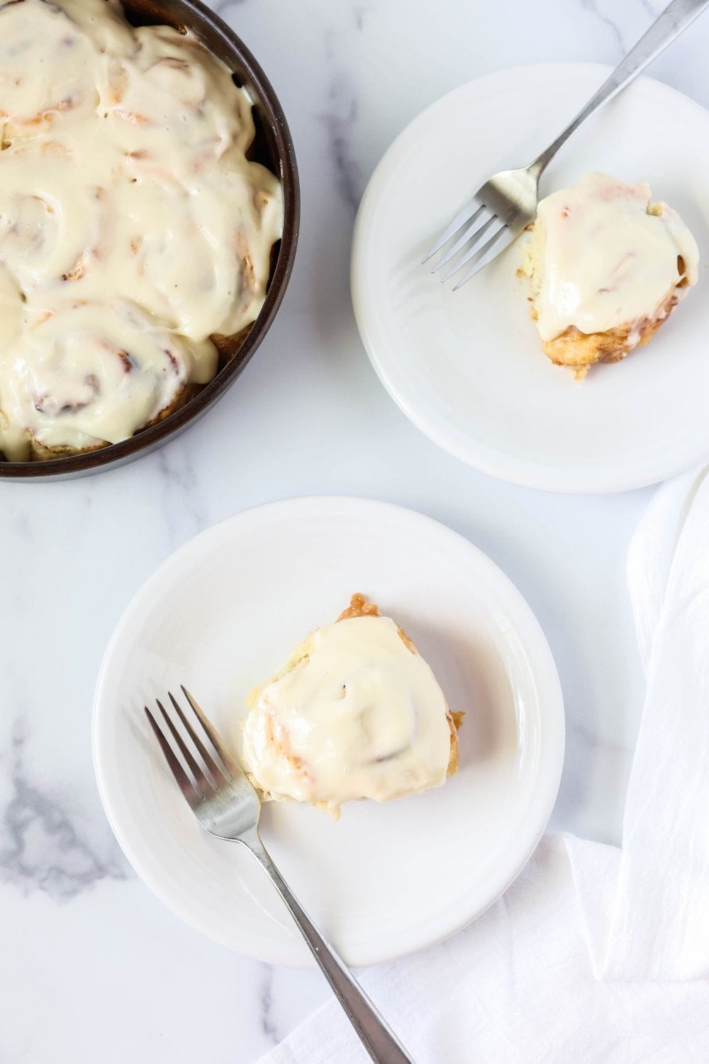 overhead view of two white plates, each with a Bisquick cinnamon roll and fork on it. The pan of cinnamon rolls is next to the plates.
