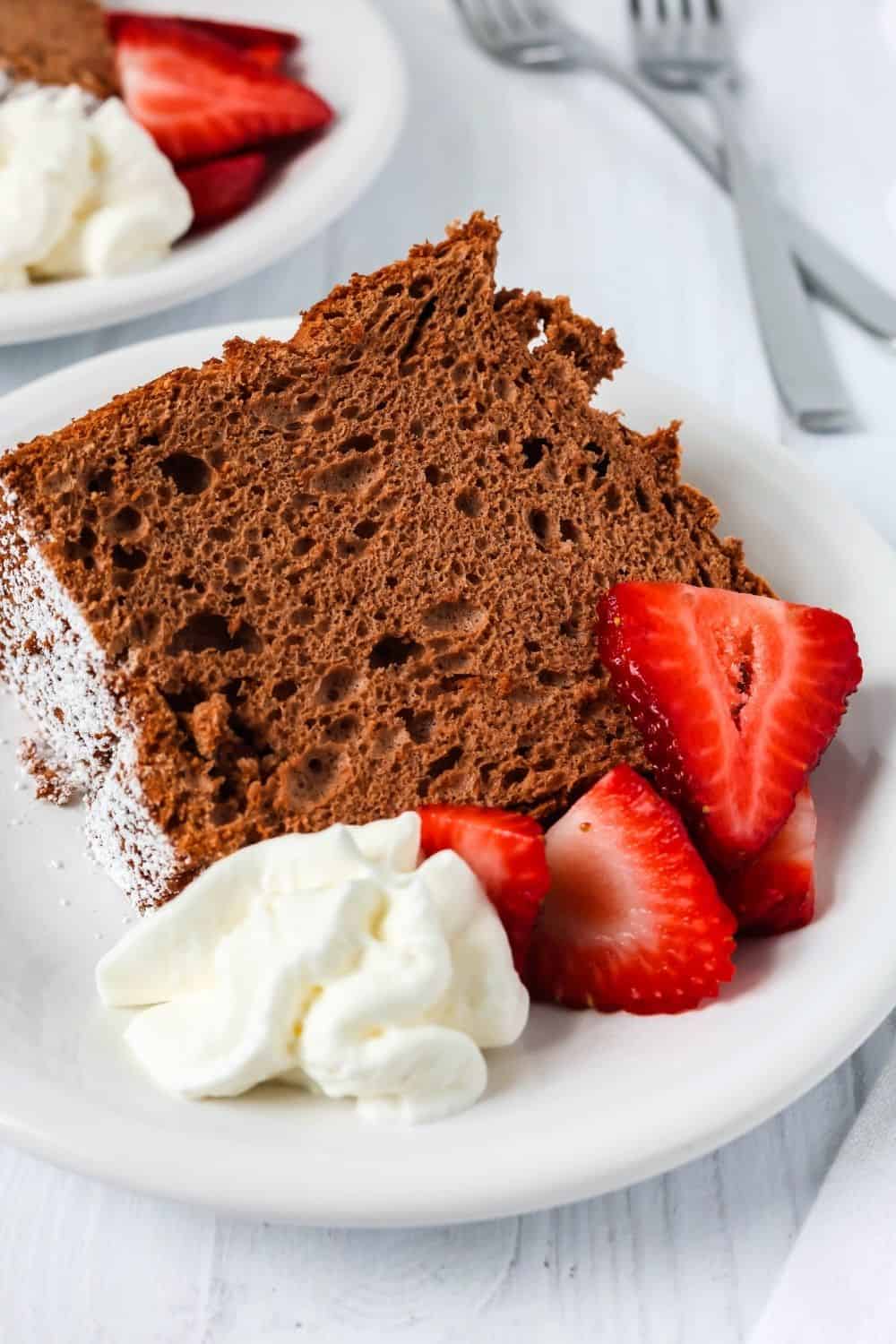 close-up view of a slice of cocoa angel food cake on a white plate, showing the light and airy interior. It's garnished with berries and cream.
