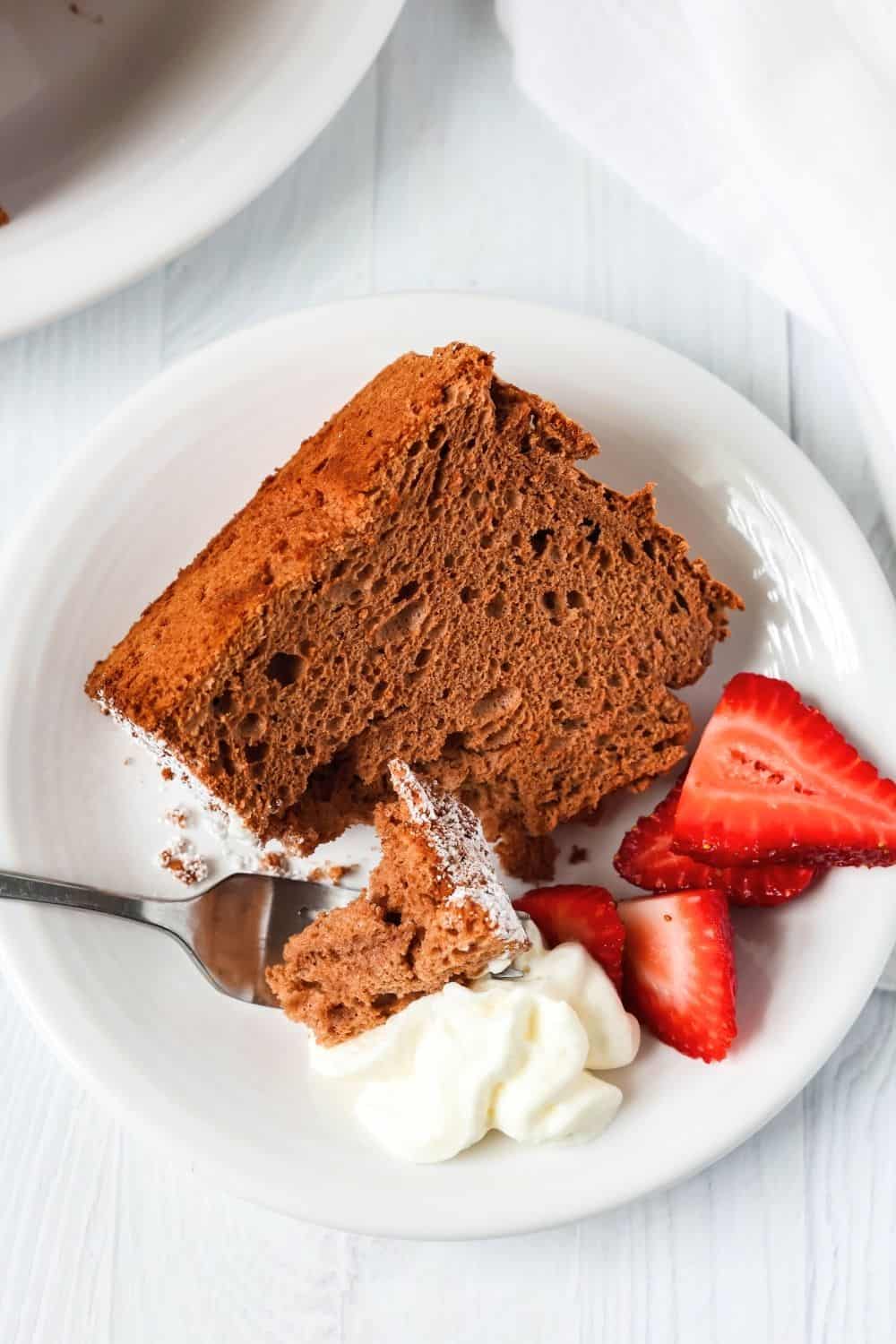 overhead view of a slice of angel food cake made with cocoa powder, on a white plate with sliced strawberries and whipped cream. A fork has taken a bite out of the cake.