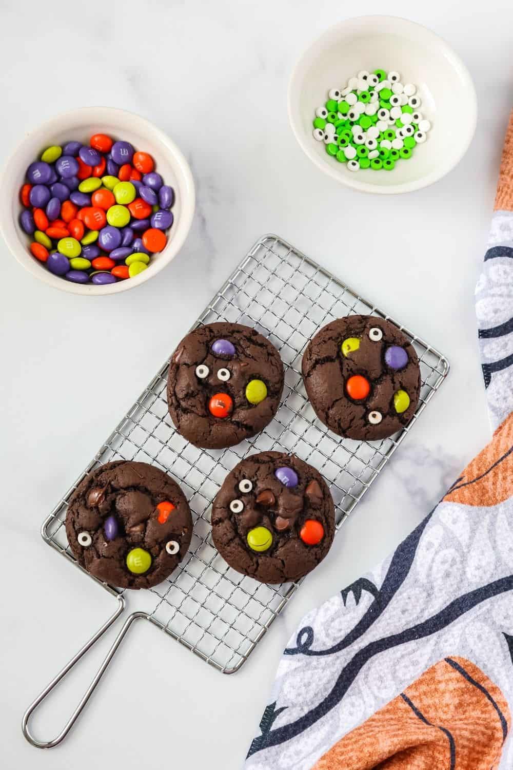 overhead view of chocolate cake mix cookies for Halloween on a wire cooling rack. Two bowls of candy eyeballs and M&Ms are next to the rack.