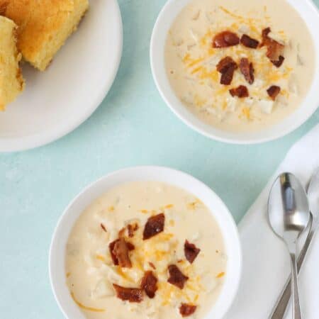 two bowls of Crock Pot hash brown potato soup, each garnished with shredded cheese and crumbled bacon. A plate of cornbread is in the background, along with two spoons.