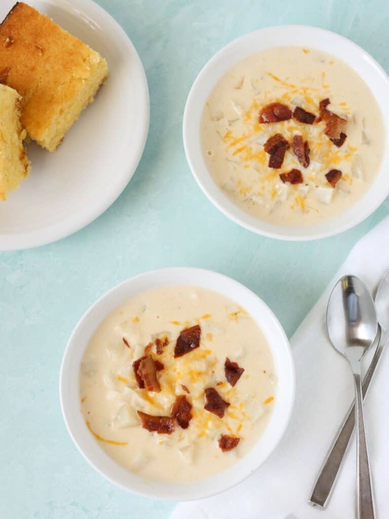 two bowls of Crock Pot hash brown potato soup, each garnished with shredded cheese and crumbled bacon. A plate of cornbread is in the background, along with two spoons.