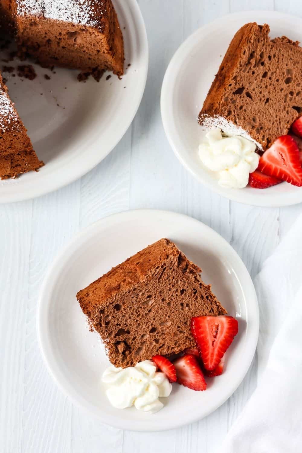 overhead view of two servings of chocolate angel food cake, each served with strawberries and cream, next to the whole tube cake.