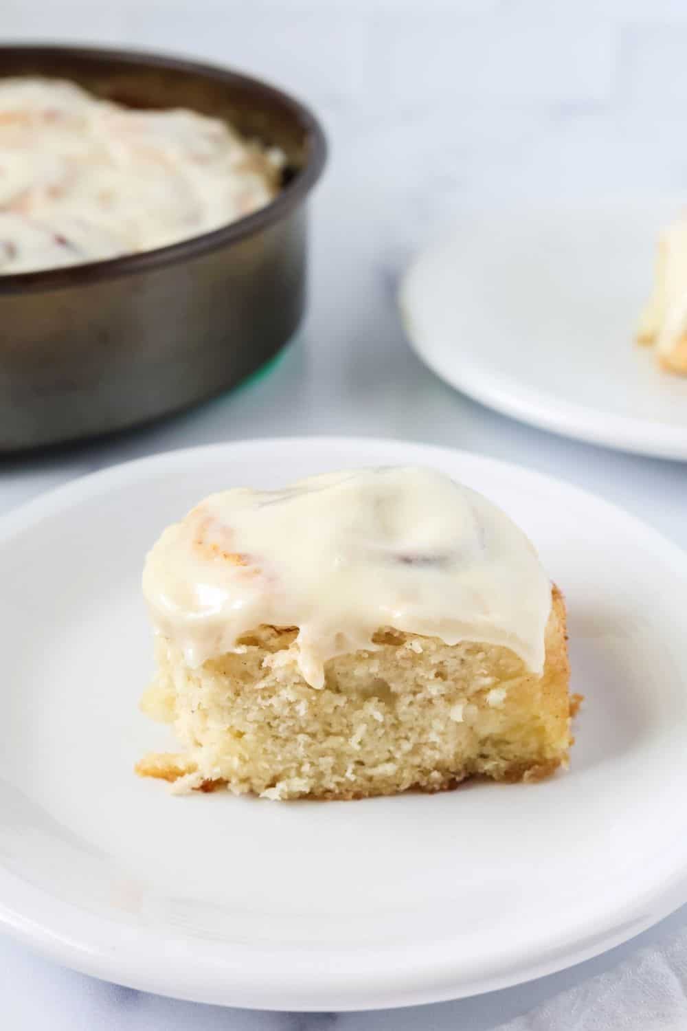 a Bisquick cinnamon bun served on a white plate. The roll is topped with cream cheese frosting, and the pan of remaining cinnamon rolls is in the background.