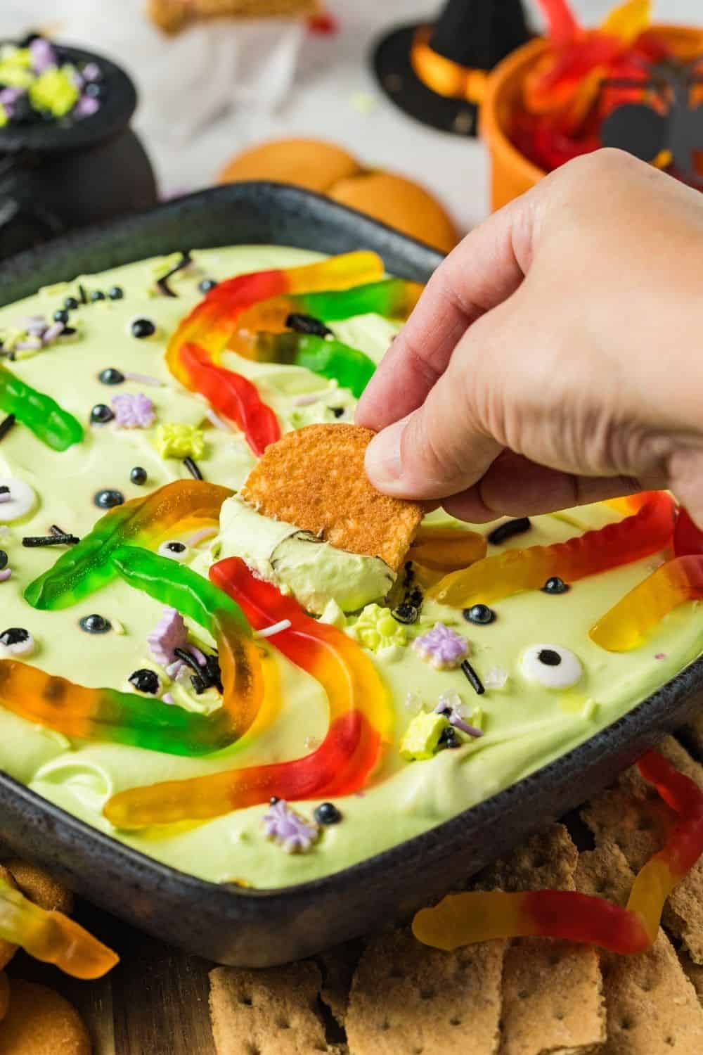 A hand dips a vanilla wafer into the cold and creamy Halloween dip.