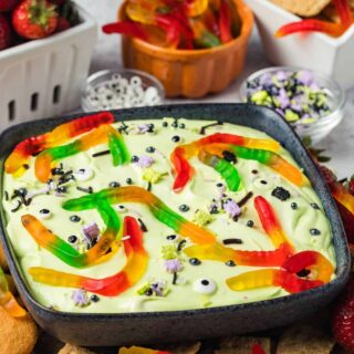 Halloween dessert dip served in a black bowl, topped with Halloween sprinkles, candy eyeballs, and gummy worms. The bowl is surrounded by strawberries, graham crackers, and vanilla wafers.