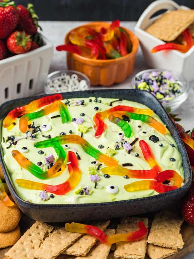 Halloween dessert dip served in a black bowl, topped with Halloween sprinkles, candy eyeballs, and gummy worms. The bowl is surrounded by strawberries, graham crackers, and vanilla wafers.