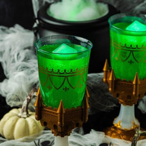 Two glasses of Witches' Brew Halloween punch served over glowing ice cubes. A steaming cauldron is in the background, and small white pumpkins and plastic spiders are scattered around the glasses of party punch.