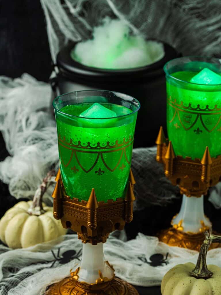 Two glasses of Witches' Brew Halloween punch served over glowing ice cubes. A steaming cauldron is in the background, and small white pumpkins and plastic spiders are scattered around the glasses of party punch.