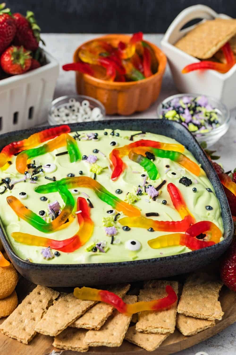side view of a black bowl filled with cold Halloween dessert dip, topped with candy eyeballs, purple and green sprinkles, and gummy worms. Surrounding the bowl of dip are garnishes and dippers.