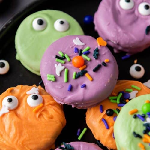 overhead view of dipped Halloween Oreos served on a black plate. Some have candy eyes pressed into the colorful candy coating, others have festive sprinkles on them.