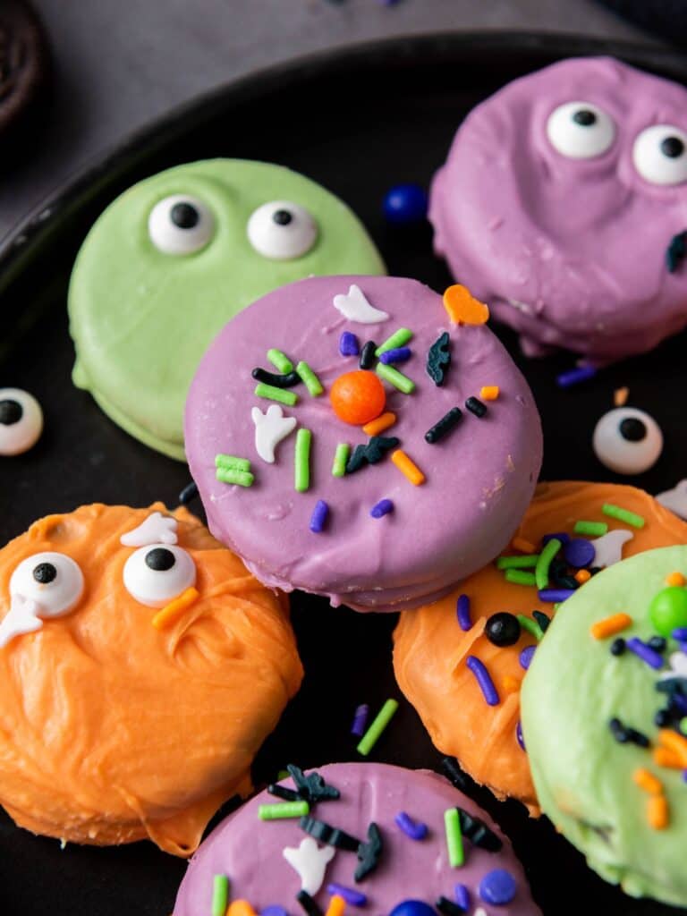 overhead view of dipped Halloween Oreos served on a black plate. Some have candy eyes pressed into the colorful candy coating, others have festive sprinkles on them.