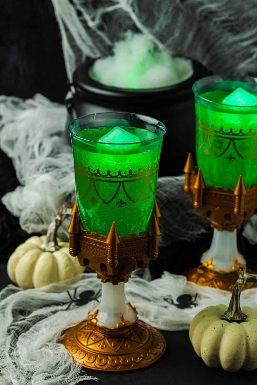 two glasses of Halloween witches' brew punch with glowing ice cubes in them. A steaming cauldron is in the background, and small white pumpkins are next to the glasses.