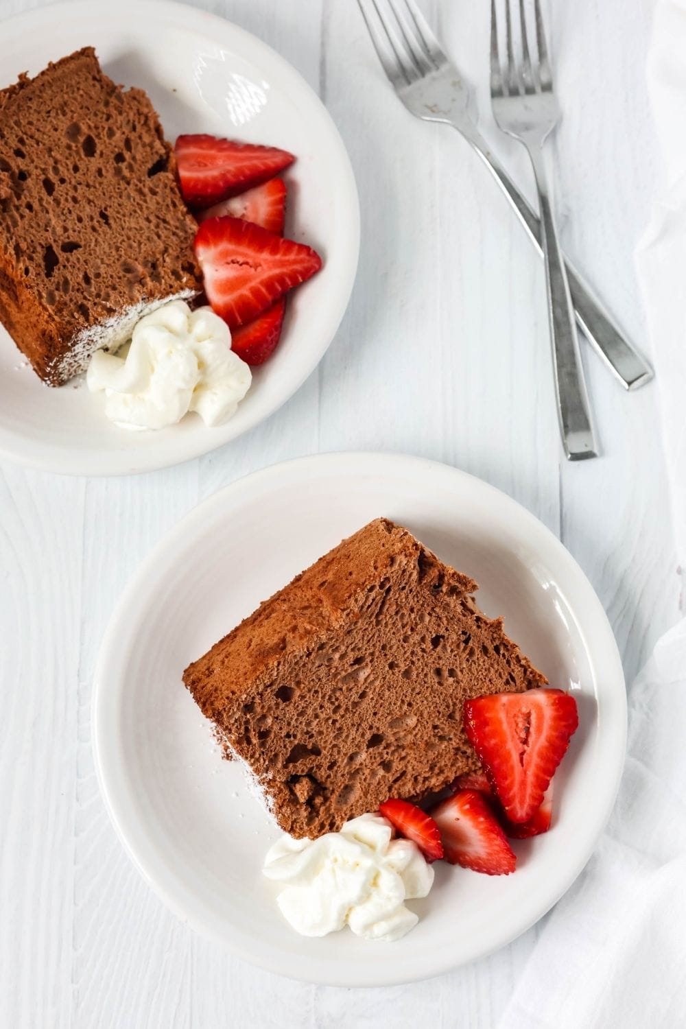 two slices of chocolate angel food cake on white dessert plates, garnished with fresh strawberries and whipped cream. Two forks are next to the plates.