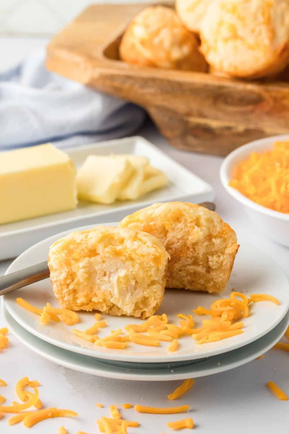 a Bisquick cheesy muffin sliced in half and spread with butter, served on a white plate. Additional muffins, butter, and cheese are in the background.