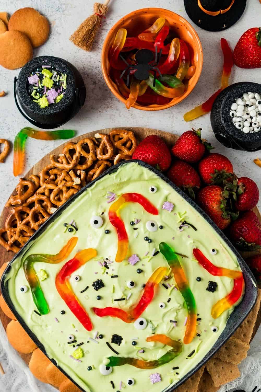 overhead view of a serving bowl filled with creamy Halloween fruit dip appetizer, topped with sprinkles and candies. Surrounding the dip bowl are pretzels, fresh strawberries, vanilla wafer cookies, and graham crackers. Sprinkles and candy eyeballs and gummy worms are near the tray.