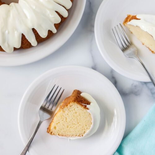 Overhead view of two slices of white mayonnaise cake served on white plates with forks. A bundt cake is in the top corner.