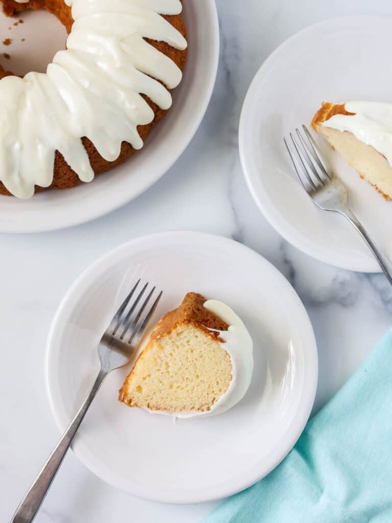 Overhead view of two slices of white mayonnaise cake served on white plates with forks. A bundt cake is in the top corner.