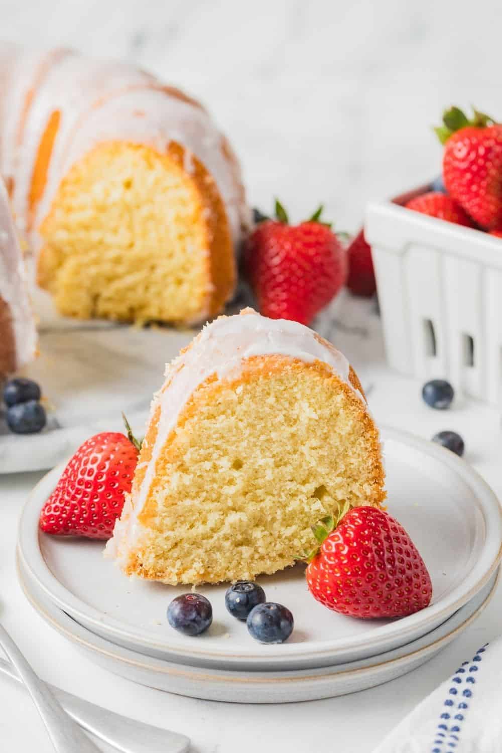 a slice of glazed vanilla pudding pound cake on a white plate with berries. The remainder of the bundt cake is in the background.