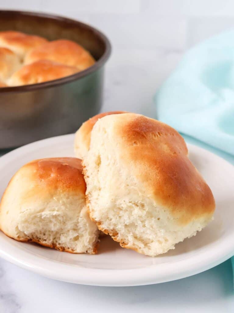 two Bisquick dinner rolls served on a white plate, with the pan of remaining yeast rolls in the background.
