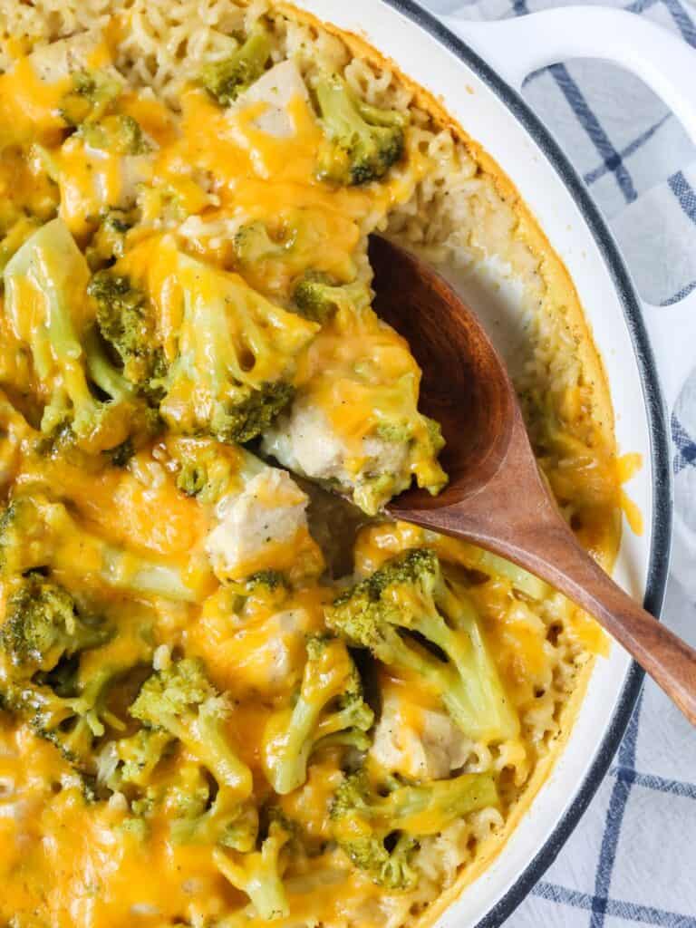 Cheesy chicken broccoli rice casserole in a Dutch oven pan. A wooden spoon is scooping out a serving of the casserole from the pan.