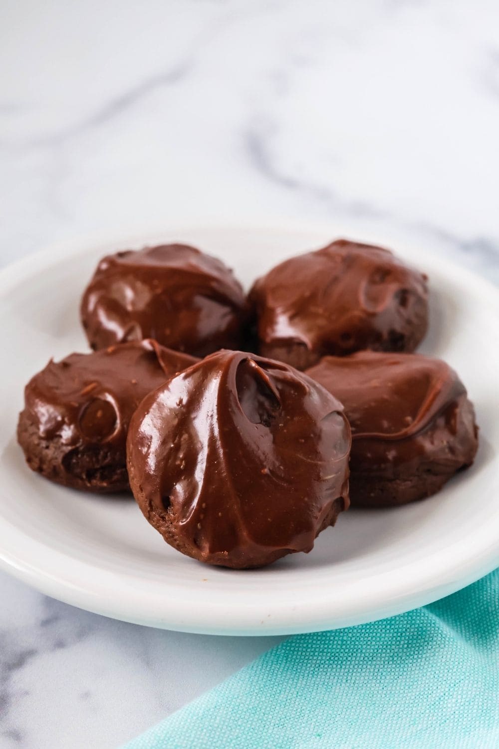 Five chocolate cookies topped with icing that hardens, served on a white plate. One cookie is propped up on its side to show the satiny finish of the frosting.