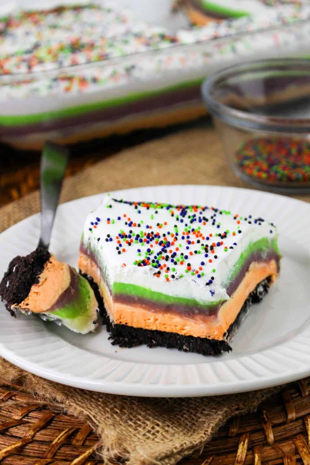 a slice of layered, no-bake, creamy Halloween lush on a white plate, with a fork holding a bite that's been cut out of it.