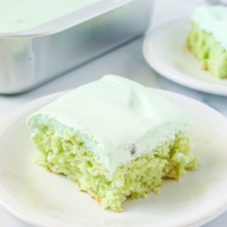 slice of pistachio pineapple cake topped with whipped cream frosting, served on a white plate. The pan of cake and another slice are in the background.