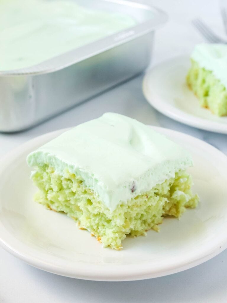 slice of pistachio pineapple cake topped with whipped cream frosting, served on a white plate. The pan of cake and another slice are in the background.