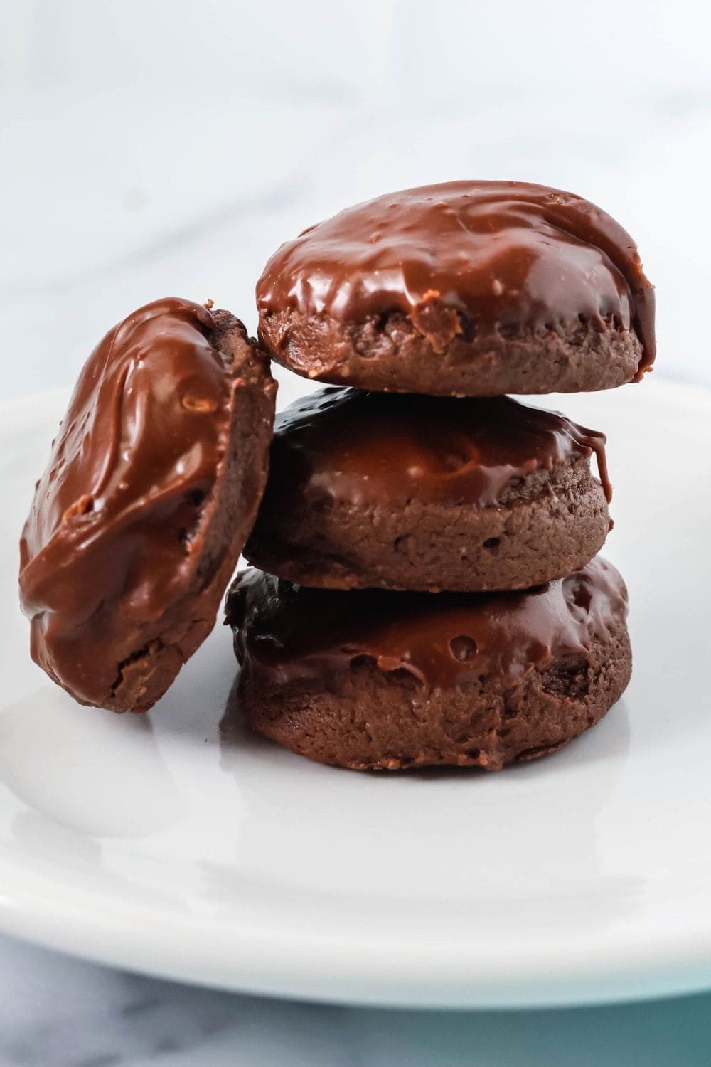 Three fudgy chocolate cookies with icing are stacked on top of each other on a white plate, with a fourth cookie propped up against the stack.