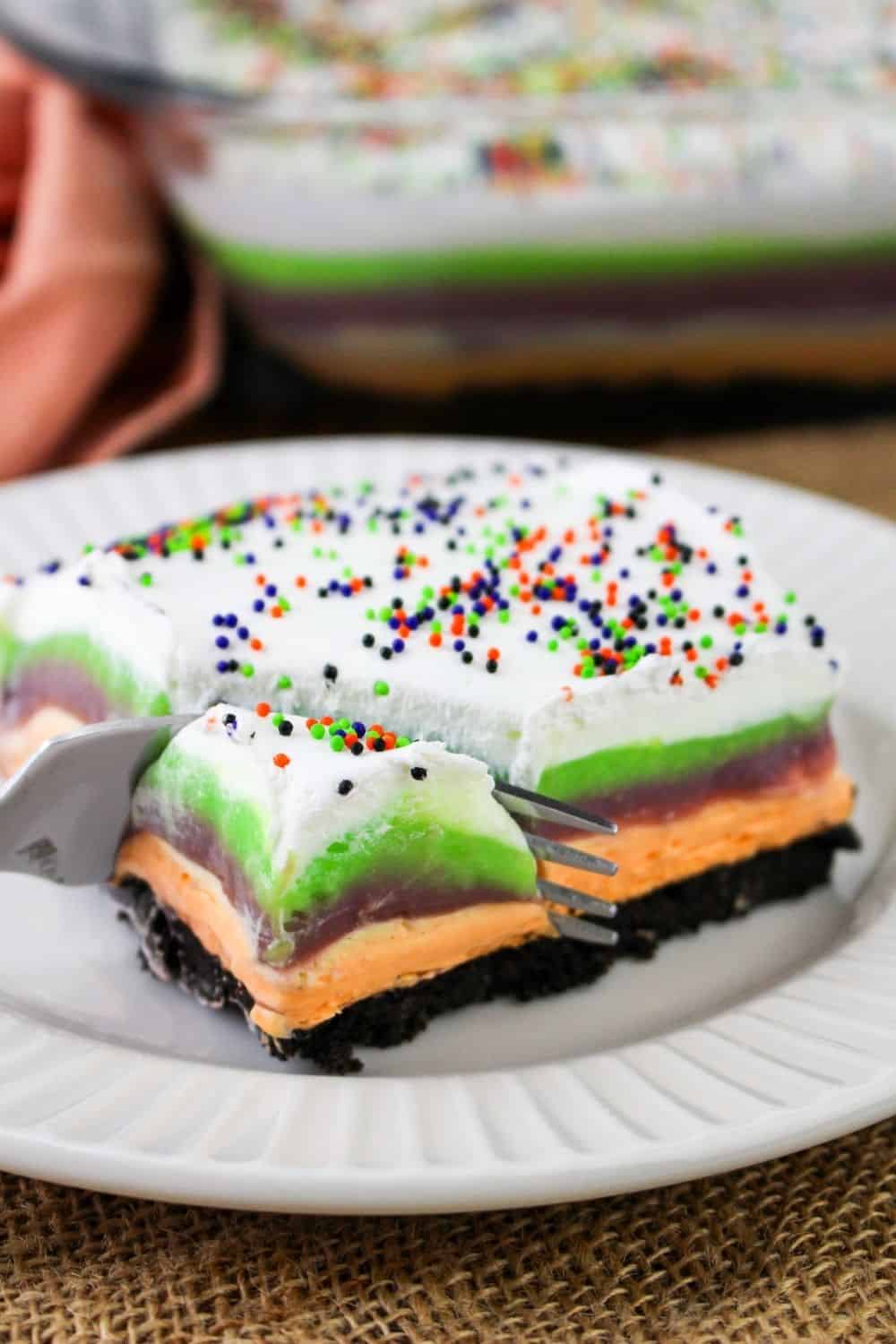 a fork slices a bite out of a square of Halloween lush dessert, made up of creamy and colorful layers.