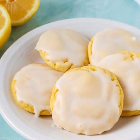 Lemon cookies made with cake mix are topped with icing and served on a white plate, with two lemon halves in the background.