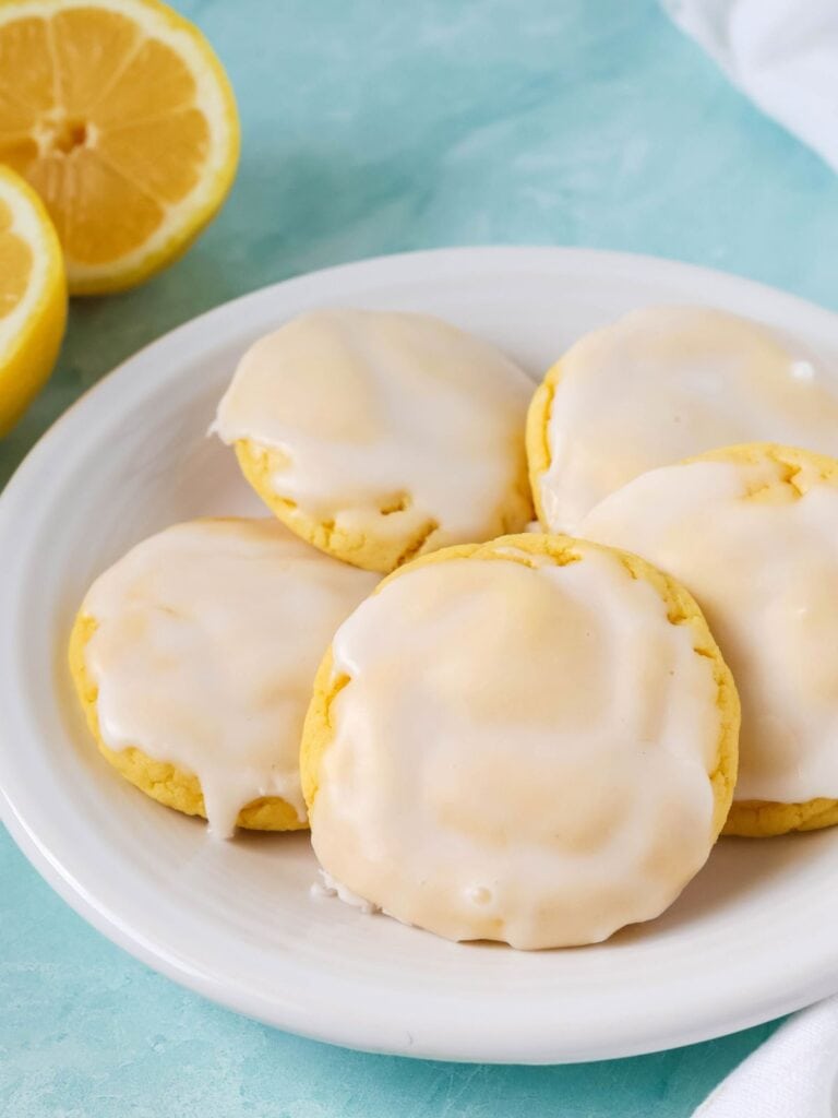 Lemon cookies made with cake mix are topped with icing and served on a white plate, with two lemon halves in the background.